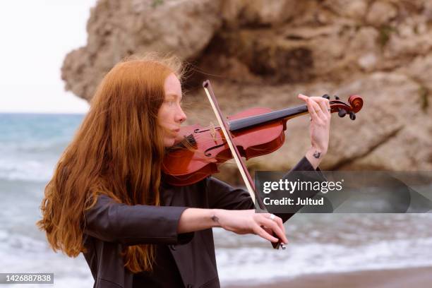 young redhead female violinist plays her violin on the beach on a cloudy day - classical music stock pictures, royalty-free photos & images