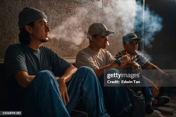 3 jóvenes asiáticos fumando sentados en fila en el callejón trasero por la noche con cigarrillo electrónico - humo de cigarrillo electrónico fotografías e imágenes de stock