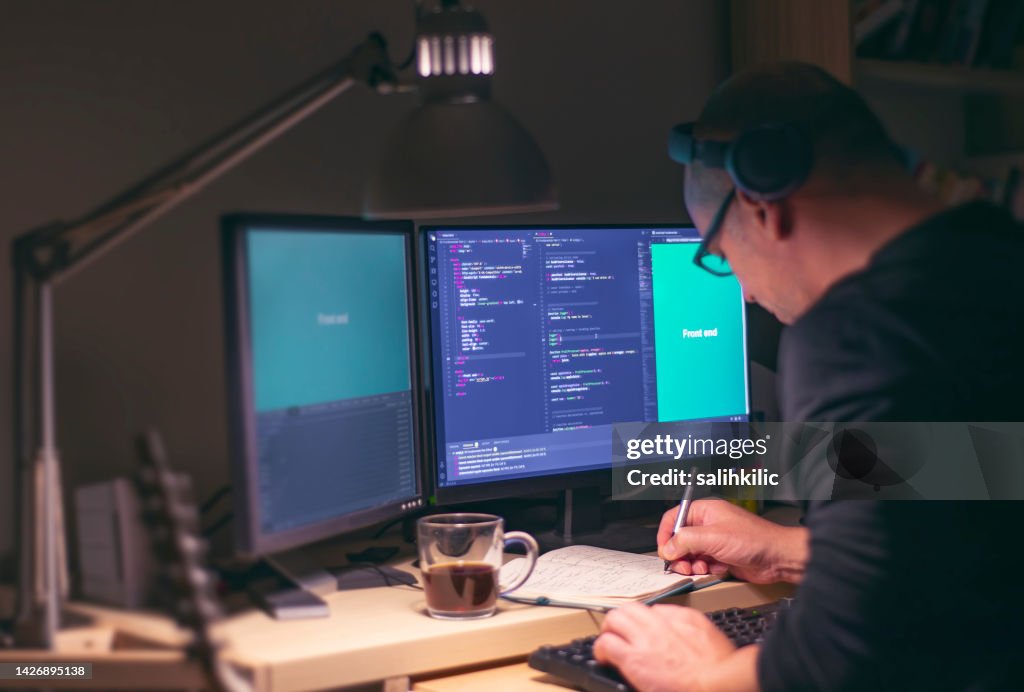 Ein männlicher Front-End-Entwickler, der Notizen auf dem Notebook macht, während er nachts programmiert