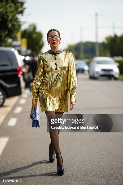 Guest wears black sunglasses, gold earrings, a gold shiny long sleeves short dress with black leather collar, black tights, black shiny varnished...