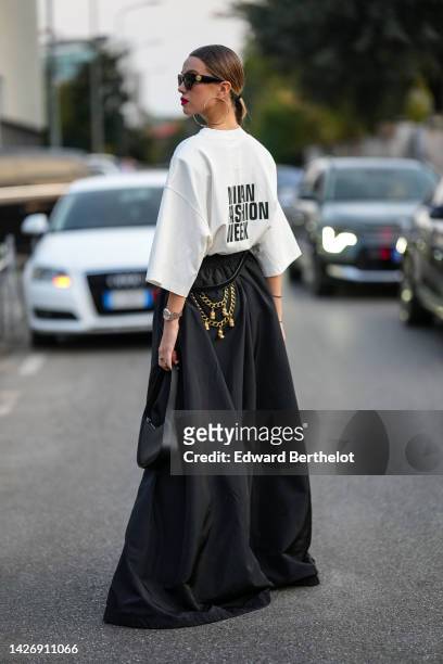 Guest wears black sunglasses from Versace, gold earrings, a white with black slogan print pattern "Milan Fashion Week" t-shirt, a black shiny leather...