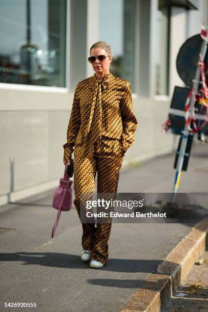 Guest wears aviator sunglasses, a yellow with brown / blue print pattern silk high neck shirt, matching yellow with brown / blue print pattern silk...