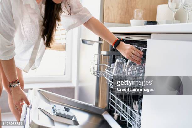 woman with long hair in white blouse puts crockery to wash in the dishwasher at home. - wasserglas stockfoto's en -beelden