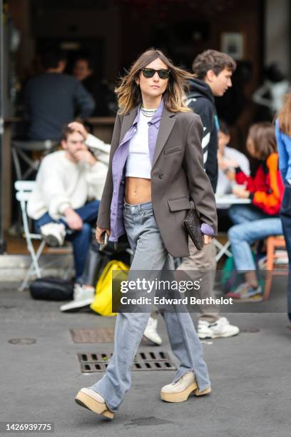 Guest wears black sunglasses, a white cropped top, a purple shirt, multicolored pearls necklaces, a gray blazer jacket, gray denim flared pants,...