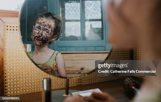 a mischievous young girl uses her mothers make-up to draw over her face, she admires her finished creation in the mirror - body paint imagens e fotografias de stock