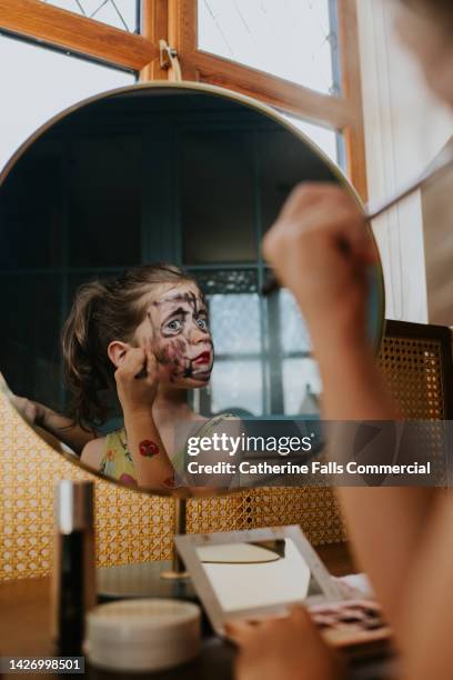 a mischievous young girl uses her mothers make-up to draw over her face, looking at herself in a mirror as she does so - 2022 a funny thing foto e immagini stock