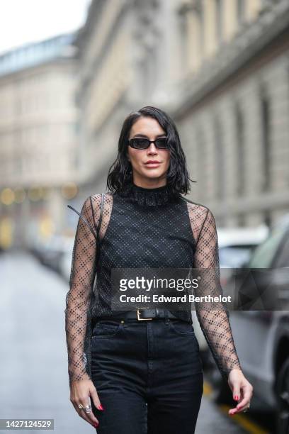 Brittany Xavier wears black sunglasses, a black high neck / sleeveless top, a black checkered print pattern transparent long sleeves blouse, a black...