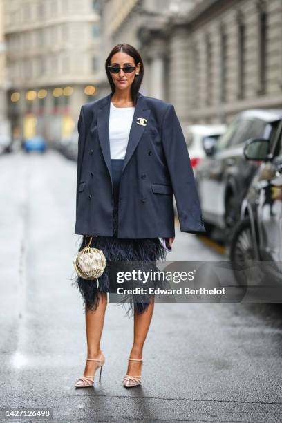 Elisa Taviti wears black sunglasses, white pearls earrings, a white t-shirt, a gray oversized blazer jacket with a gold and rhinestones logo brooch...