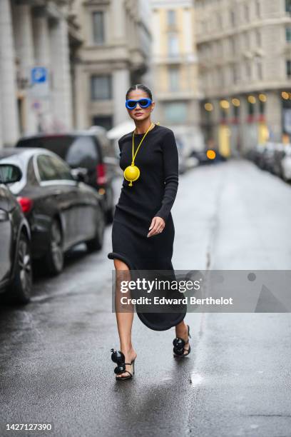 Thassia Naves wears blue puffy sunglasses from Loewe, a black long sleeves / asymmetric rolled-up knees dress, a yellow shiny leather balloon pendant...