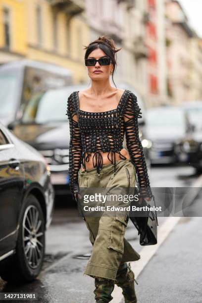 Angela Rozas Saiz wears black sunglasses, gold logo earrings from Dolce & Gabbana, a black shiny leather laces square-neck / long sleeves cropped top...