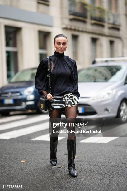 Mary Leest wears gold pendant logo earrings from Dolce & Gabbana, a black turtleneck / puffy long sleeves blouse, a black shiny leather shoulder bag...