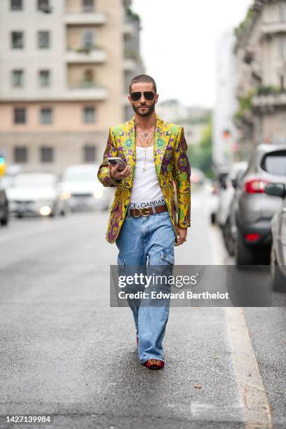 Pelayo Diaz wears black sunglasses, gold long necklaces, a white tank-top, a yellow gold with embroidered green / blue / pink flower pattern blazer...