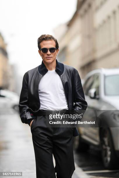 Carlo Sestini wears black sunglasses, a white t-shirt, a black shiny leather bomber coat, a black shiny leather belt, black suit pants, boutside...