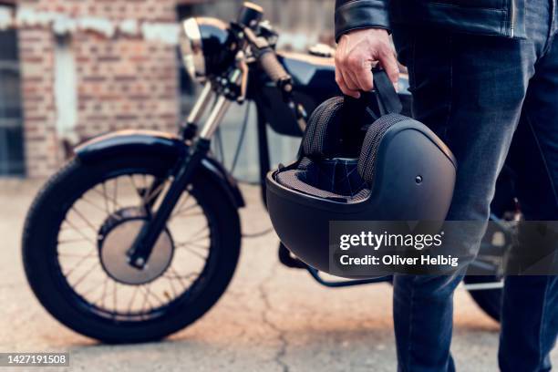 unrecognizable motorcyclist carries his motorcycle helmet in one hand. in the background is his parked motorcycle - biker stockfoto's en -beelden