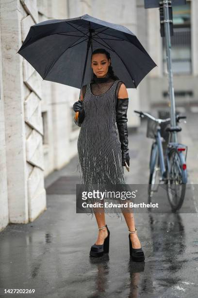 Guest wears a black large umbrella, silver earrings, a black mesh / fishnet turtleneck sleeveless top, a black with embroidered silver rhinestones...