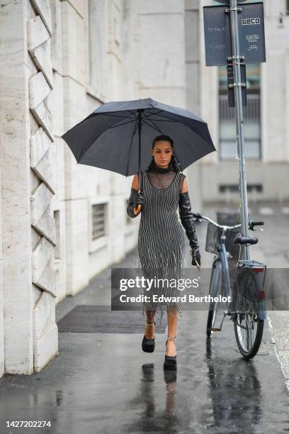 Guest wears a black large umbrella, silver earrings, a black mesh / fishnet turtleneck sleeveless top, a black with embroidered silver rhinestones...