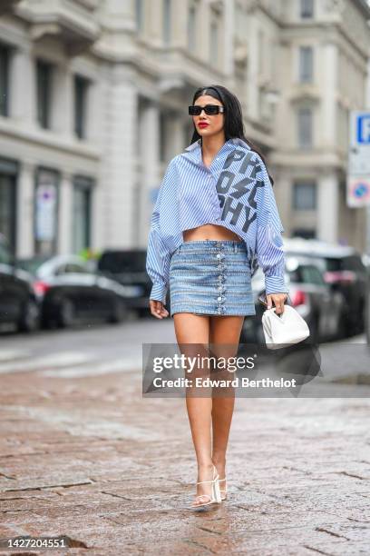 Guest wears black sunglasses, a navy blue and white striped print pattern with black large embroidered logo cropped shirt from Philosophy, blue faded...