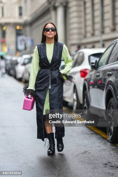 Guest wears black futurist sunglasses, a pale green long sleeves / midi dress with embroidered black feathers borders, a black long nylon sleeveless...