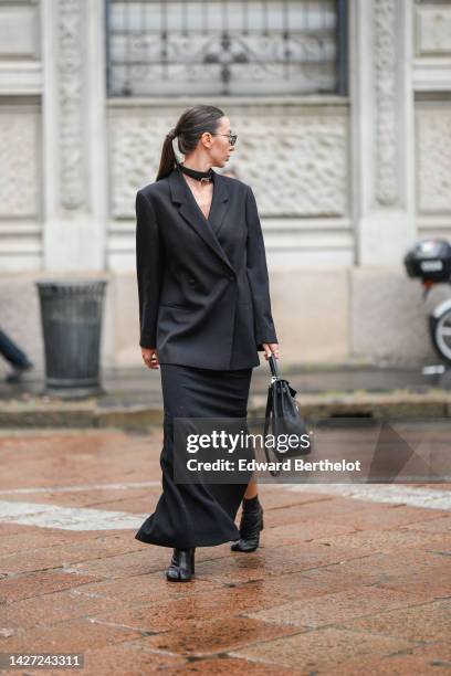 Guest wears black sunglasses, a black shiny leather Kelly belt from Hermes as a necklace, a black blazer jacket, a black long dress, a black shiny...