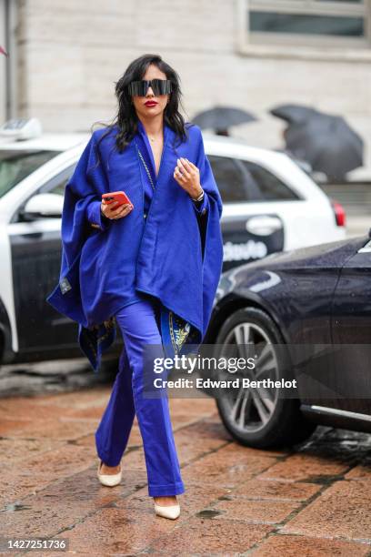 Guest wears black futurist sunglasses, a royal blue oversized denim cloak, a royal blue blazer jacket, royal blue suit pants, silver and gold rings,...