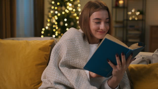 Cheerful girl with brown hair sits on the couch and reads book in blue binding. The girl reads funny story and smiles. She has black nails and is covered with plaid