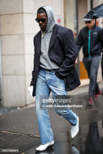Guest wears a pale gray hoodie sweater, a black blazer jacket, a black shiny leather belt , blue denim pants, white shiny leather pointed / block...