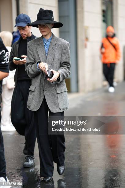 Guest wears a black large hat, a blue denim shirt, a gray blazer jacket, black flared pants, black shiny leather pointed ankle boots , outside...