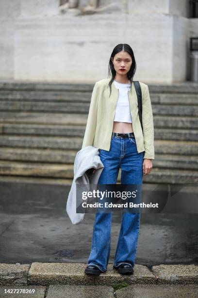 Guest wears a white cropped top, a white latte jacket, a black shiny leather shoulder bag, a black shiny leather belt, blue denim flared pants, a...