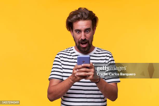 handsome blonde man standing by a yellow background holding a mobile surprised by what he sees in it. - hombre asombrado fotografías e imágenes de stock