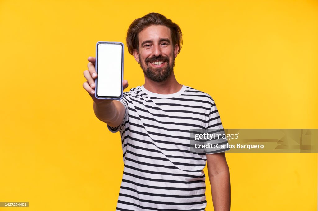 Cheerful photo of a caucasian young man showing his phone with blank space on camera and smiling standing in an isolated yellow background