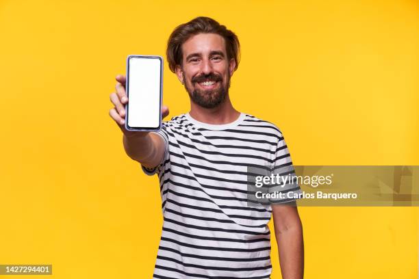 cheerful photo of a caucasian young man showing his phone with blank space on camera and smiling standing in an isolated yellow background - halten stock-fotos und bilder