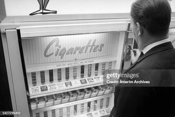 View of a man purchasing cigarettes at a cigarette vending machine, April 1965.