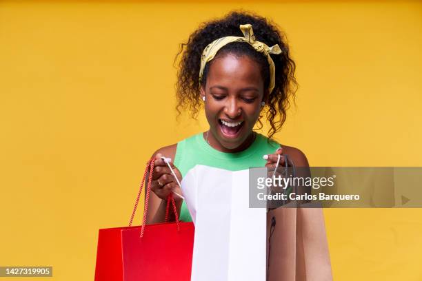 cheerful latin american young woman opening birthday gifts. isolated in a yellow background. concept of big sales. - latin american and hispanic shopping bags stockfoto's en -beelden