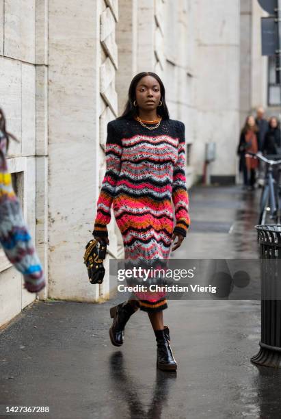 Coco Bassey wears long knitted dress in black red white grey tones and long sleeves, black studded bag, black ankle boots with zipper, necklace in...