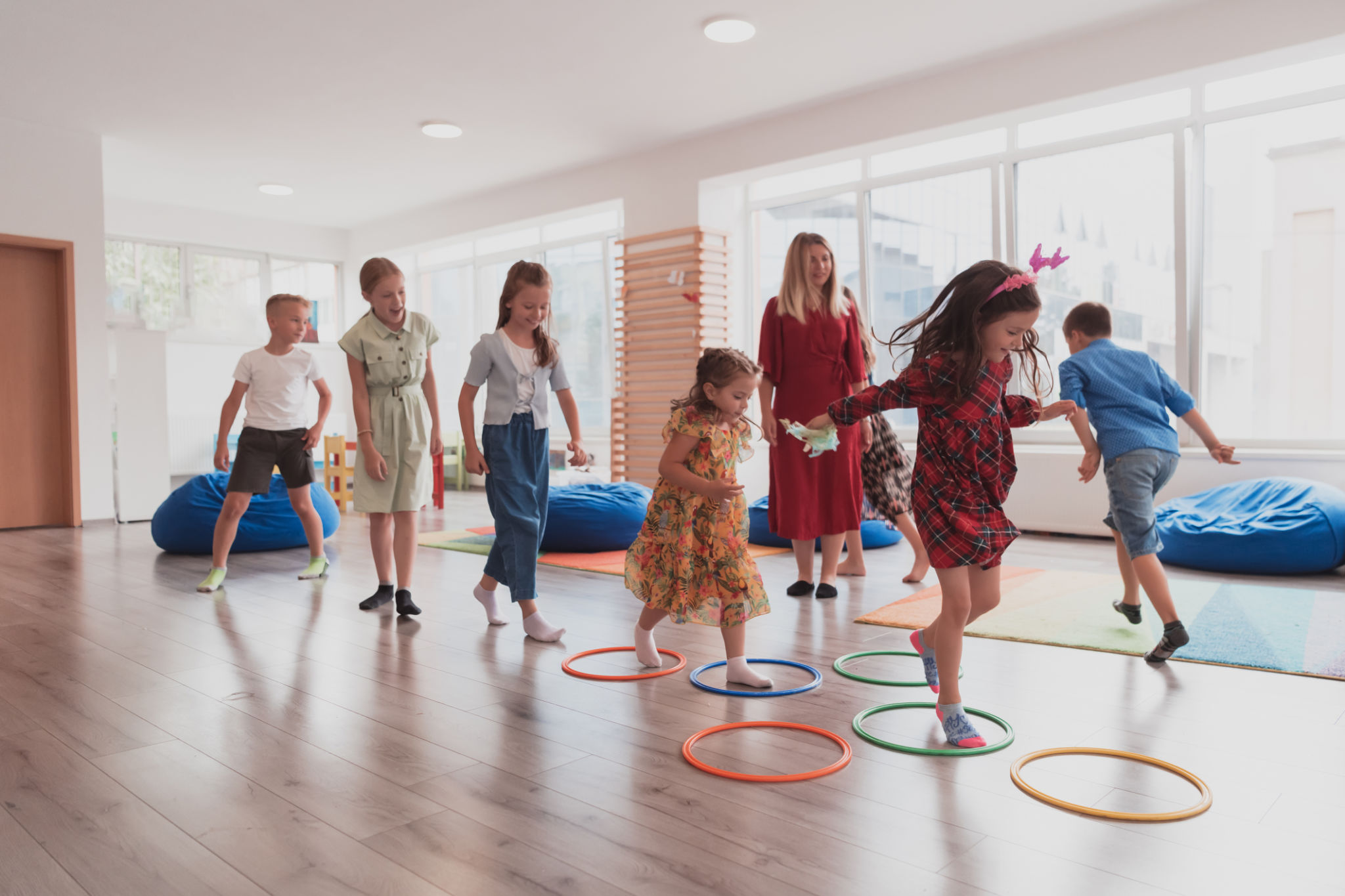 Small nursery school children with female teacher on floor indoors in classroom, doing exercise. Jumping over hula hoop circles track on the floor. Small nursery school children with female teacher on floor indoors in classroom, doing exercise. Jumping over hula hoop circles track on the floor.