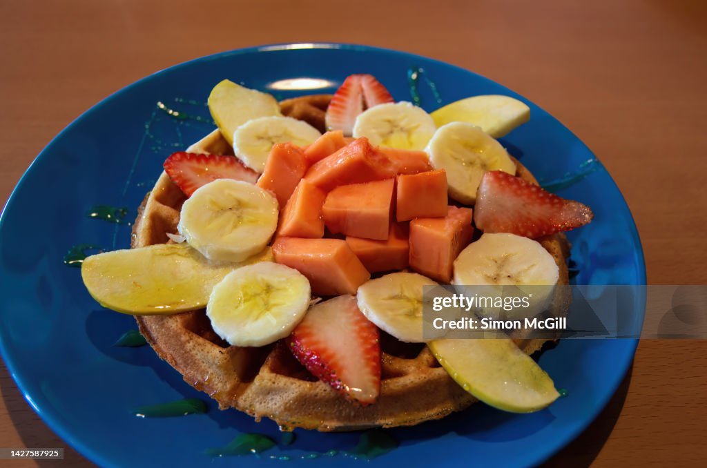 Waffle with chopped apple, papaya, banana and strawberry , drizzled with honey, on a blue plate on a cafe table