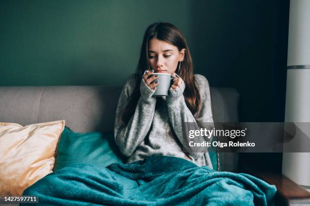 cold sick season, trying to get some warm. sick young woman drink hot tea, wrapped in blanket in cold apartment - winter illness stock pictures, royalty-free photos & images