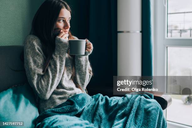 depression and frustration at home. young shivering woman drink hot tea, looking out the window, wrapped in a blanket in cold apartment - beef stockfoto's en -beelden