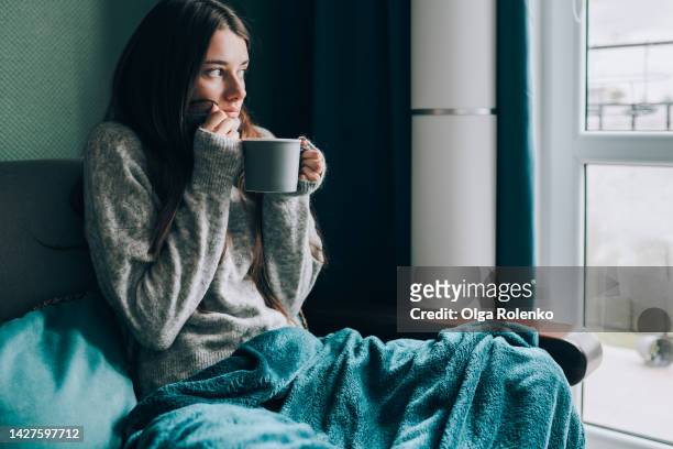 depression and frustration at home. young shivering woman drink hot tea, looking out the window, wrapped in a blanket in cold apartment - wrapped in a blanket stock pictures, royalty-free photos & images