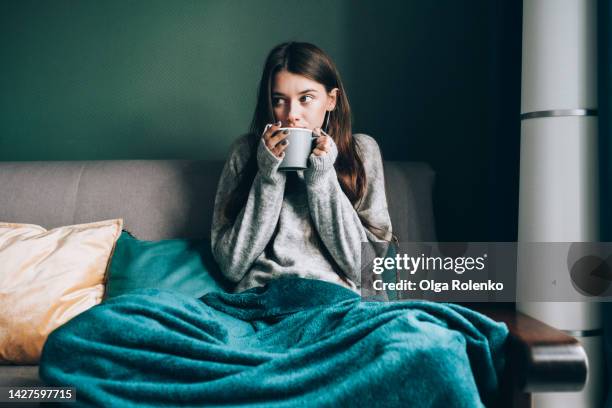 sick leave and seasonal indisposition at home. young woman drink hot tea, wrapped in blanket in cold apartment - flu stockfoto's en -beelden