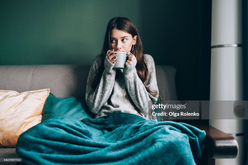 Sick leave and seasonal indisposition at home. Young woman drink hot tea, wrapped in blanket in cold apartment