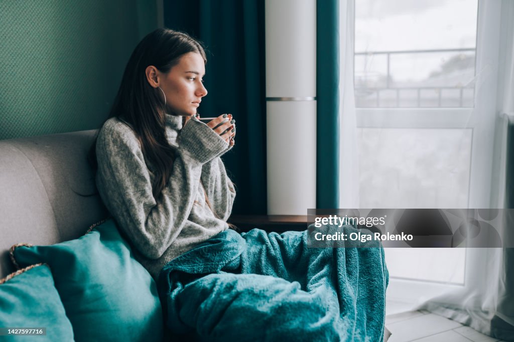 No central heating, cold apartment. Young brunette woman drink hot tea, look out the window, wrapped in a blanket in cold apartment