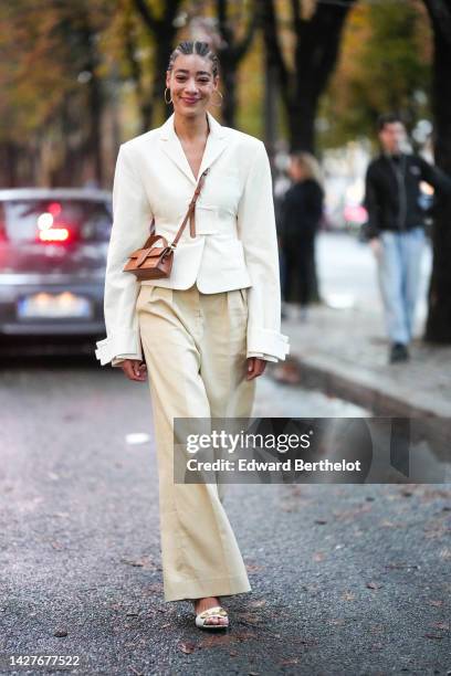 Guest wears earrings, a white blazer jacket, a brown Jacquemus bag, beige flare pants, outside Jacquemus, during Paris Fashion Week - Womenswear...