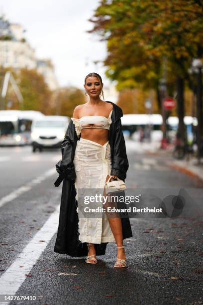 Veronica Ferraro wears earrings, a black long coat, a white crop top, a white slit skirt, a Jacquemus bag, sandals, outside Jacquemus, during Paris...