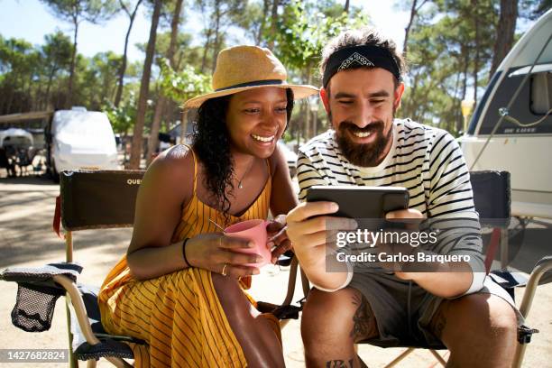 young multiracial couple sharing smart phone while sitting on camping chairs with caravans at the background enjoying their holidays. - camping chair stock pictures, royalty-free photos & images