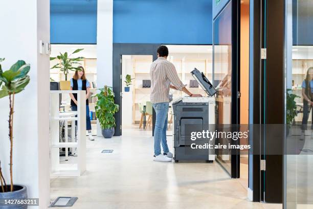 man in jeans standing using a printer next to a glass wall while talking to his coworker - プリンター ストックフォトと画像