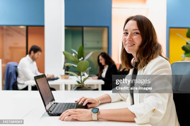 long-haired woman looking and smiling at the camera, sitting at her desk using a laptop, two coworkers and a glass wall in the background. - internship marketing stock-fotos und bilder