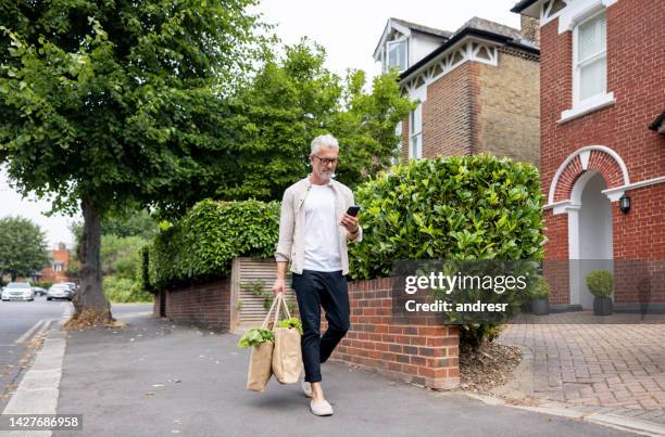 man walking home with the groceries and using his cell phone - mature men walking stock pictures, royalty-free photos & images