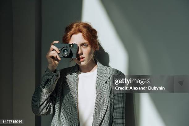 beautiful young redhead woman holding a camera in her hand looking at the camera indoors. copy space - paparazzi photographers stock-fotos und bilder