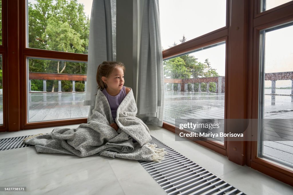 A girl in a plaid is sitting at a large window and looking at the rain.